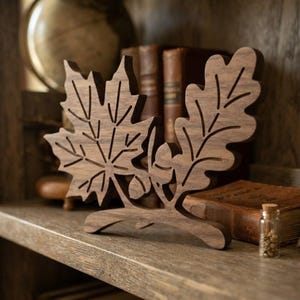 May include: A dark brown wooden decorative cutout featuring a maple leaf and an oak leaf attached to a single branch. The design includes two small acorns nestled between the leaves, showcasing a natural wood grain texture and precise laser-cut details.