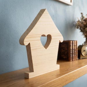 May include: A light-colored wooden house-shaped decorative object featuring a cutout heart shape in the center. The item has a flat base for stability and displays a natural wood grain pattern throughout its smooth, light brown surface.