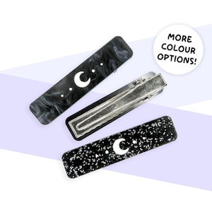 May include: Three rectangular hair clips with celestial designs. One clip is black with a white crescent moon and dots, another is black and white marbled, and the third is black with white speckles and a crescent moon. The text "MORE COLOUR OPTIONS!" is in a white circle.