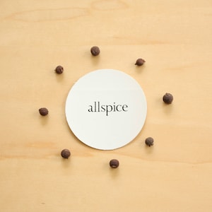 May include: A white circle with the word "allspice" printed in black on a light wood background. There are eight small, dark brown peppercorns arranged around the circle.