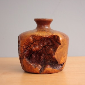 Puede incluir: Un jarrón de madera hecho a mano con una estética natural y orgánica. El jarrón presenta un cuerpo bulboso y un cuello estrecho, con un color marrón cálido. La veta y los nudos de la madera son visibles, lo que añade a su carácter único.