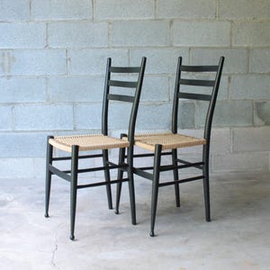 May include: Two black wooden chairs with woven seats. The chairs have a ladder back design and tapered legs. The seats are a light tan color, contrasting with the black frames. The chairs are positioned in front of a gray cinder block wall.