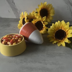 May include: A yellow ceramic bowl filled with orange, yellow, and white candy corn, accompanied by a smooth, conical ceramic object colored in orange and white, and several bright yellow artificial sunflowers with dark brown centers.