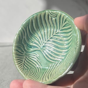 May include: A small, round, light green ceramic dish featuring a debossed fern leaf pattern across its surface. The glaze has a glossy finish with subtle variations in color depth, highlighting the intricate texture of the pressed botanical design.