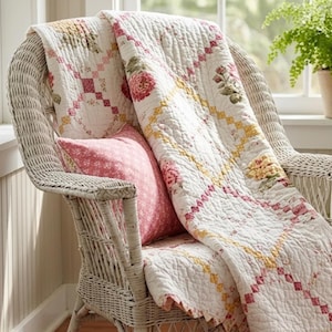 Puede incluir: Una silla de mimbre blanca sostiene una manta acolchada y un cojín rosa. La manta presenta un patrón de diamantes con motivos florales en tonos rosa, amarillo y verde. El cojín tiene un estampado floral blanco pequeño sobre fondo rosa.
