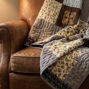 May include: A patchwork quilt featuring alternating squares of solid grey fabric and cream fabric printed with a pattern of brown, green, and burgundy leaves. The quilt has a visible quilted texture and is draped over a brown leather armchair.