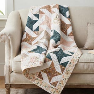 May include: A quilted throw blanket featuring a geometric star pattern in shades of dark teal, muted peach, and white. The fabric displays a speckled texture throughout, with a floral-patterned border along the edges of the rectangular textile.