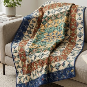 May include: A quilted throw blanket featuring a geometric pattern of triangles in shades of rust orange, teal, mustard yellow, and cream. The blanket is finished with a dark blue fabric border and visible stitching throughout the surface.