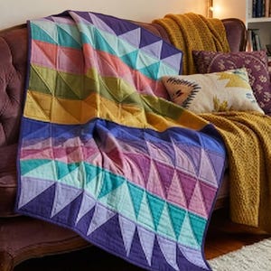 Puede incluir: Una manta acolchada con un patrón geométrico de triángulos en tonos morado, turquesa, rosa, amarillo y azul claro. El tejido muestra un diseño triangular repetitivo con líneas de costura visibles en toda la superficie de la tela.