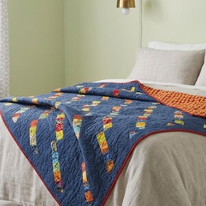 May include: A blue quilt featuring a pattern of vertical, multi-colored fabric strips with a textured, wavy stitching design. The quilt has a thin red border and a contrasting orange, textured underside that is visible at the folded corner.