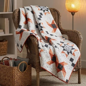 May include: A white quilt featuring a geometric star pattern in shades of burnt orange and charcoal grey. The fabric displays a textured, quilted surface with intricate stitching throughout, draped over a woven wicker chair with a rust-coloured trim.