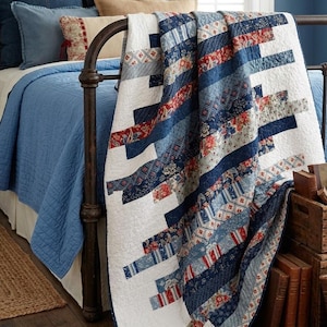 May include: A quilt featuring a diagonal striped pattern of various blue, white, and red fabrics with floral, plaid, and geometric prints. The quilt has a white background and is draped over a dark metal bed frame, showcasing a quilted texture.