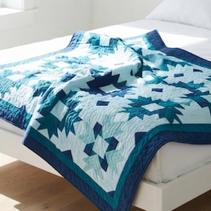 May include: A quilted bedspread featuring a geometric star pattern in shades of navy blue, teal, and light blue. The fabric has a visible stitched texture throughout, with a solid dark blue border framing the intricate, repeating star design.