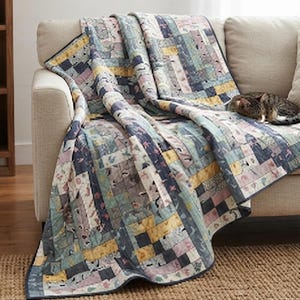 May include: A patchwork quilt featuring a rectangular block pattern in muted shades of blue, yellow, pink, and grey. The fabric displays various small-scale floral and geometric prints. A tabby cat is resting on the folded fabric surface.