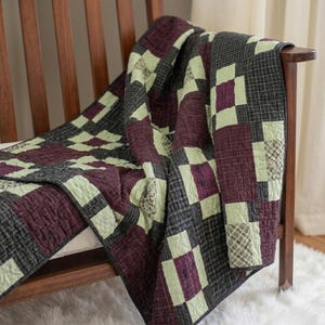 Peut inclure: Une courtepointe en patchwork présentant un motif de blocs répétitifs dans des tons de prune profond, de gris anthracite et de vert sauge clair. Les carrés de tissu affichent un mélange de couleurs unies, de petits carreaux et de textures.