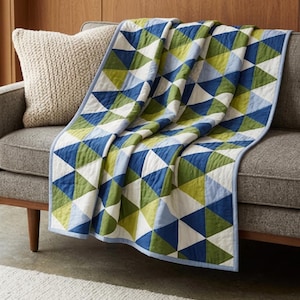 May include: A quilted throw blanket featuring a geometric pattern of triangles in shades of navy blue, olive green, light blue, and white. The blanket has a light blue fabric border and is draped over a gray sofa with a textured cream pillow.