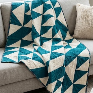 May include: A quilt featuring a geometric pattern of alternating teal and off-white triangles. The fabric displays visible linear stitching throughout the surface, creating a textured appearance. The edges are finished with a dark patterned binding.