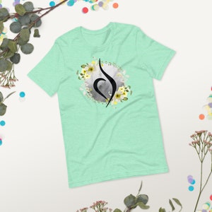 May include: A light green t-shirt featuring a black and white floral design. The design incorporates a stylised symbol with a heart shape and a circle around it. The symbol is surrounded by white flowers with yellow centres.