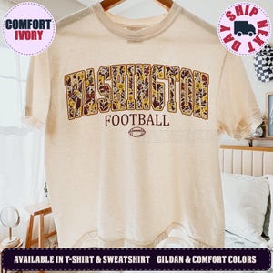May include: An ivory short-sleeve t-shirt featuring the word WASHINGTON in a block font filled with a floral pattern in shades of brown, yellow, and white. Below the text is the word FOOTBALL and a small brown graphic of a football.