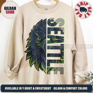 May include: A sand-colored crewneck sweatshirt featuring a graphic of a stylized eagle head in shades of dark blue and bright green. The word SEATTLE is printed vertically in large, bold, grey block letters next to the eagle illustration.
