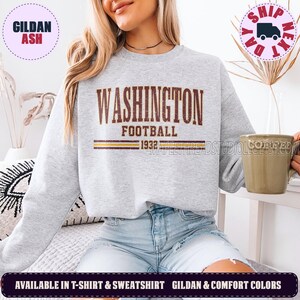 May include: A light grey crewneck sweatshirt featuring the text WASHINGTON FOOTBALL 1932 in a distressed brown font. The design includes two horizontal lines in brown and yellow below the text. The garment has long sleeves and a ribbed neckline.