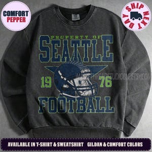 May include: A charcoal grey crewneck sweatshirt featuring a distressed graphic print. The text reads PROPERTY OF SEATTLE FOOTBALL with the numbers 19 and 76 flanking a central dark blue football helmet illustration set against a large star.