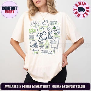 May include: An ivory t-shirt featuring a graphic print with the text Let's go Seattle, Emerald City, 12s, 50, 206, Game Day, Loud and Proud, and #1. The design includes illustrations of a football, eagle, helmet, foam finger, and goal post.
