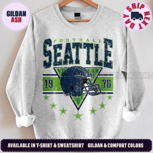 May include: A light gray crewneck sweatshirt featuring a distressed graphic with the text SEATTLE FOOTBALL 1976. The design includes a dark blue football helmet centered over a green triangle, surrounded by small green stars on a light gray background.