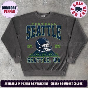 May include: A charcoal grey crewneck sweatshirt featuring a graphic of a dark blue football helmet centred above a green football field graphic. The text reads FOOTBALL SEATTLE EST. 1976 SEATTLE, WA in dark blue and green collegiate lettering.
