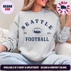 May include: A light grey crewneck sweatshirt featuring dark blue text that reads SEATTLE FOOTBALL with EST. 1976. Centered between the words is a graphic of a football containing a silhouette of the Seattle skyline including the Space Needle.