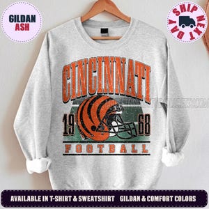 Op de afbeelding: Een lichtgrijs sweatshirt met ronde hals en een verweerde afbeelding van een oranje en zwart gestreepte footballhelm. De tekst CINCINNATI, 1968 en FOOTBALL staat in oranje blokletters op het shirt, inclusief horizontale groene strepen.