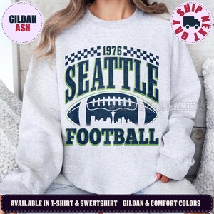 May include: A light grey crewneck sweatshirt featuring a dark blue and green graphic print. The design includes the text 1976 SEATTLE FOOTBALL, a checkered border, and a football illustration containing a silhouette of the Seattle city skyline.