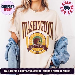 May include: An ivory t-shirt featuring a graphic print with the text WASHINGTON in large maroon letters above a circular emblem. The emblem includes the words ESTABLISHED FOOTBALL 1932, a maroon football helmet, and a small football icon.