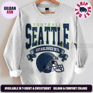 May include: A light grey crewneck sweatshirt featuring a dark blue and green graphic print. The design includes the text FOOTBALL SEATTLE ESTABLISHED 1976 and a dark blue football helmet illustration with a distressed, vintage-style texture.