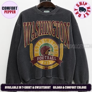May include: A charcoal grey crewneck sweatshirt featuring the word WASHINGTON in large maroon letters. Below, a circular graphic displays a maroon football helmet, the text ESTABLISHED FOOTBALL 1932, and a small football icon in gold.
