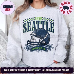 May include: A light gray crewneck sweatshirt featuring a dark blue football helmet graphic with the text SEATTLE and 1976 above it. A checkered pattern borders the top, and a banner with the word FOOTBALL sits below the helmet design.
