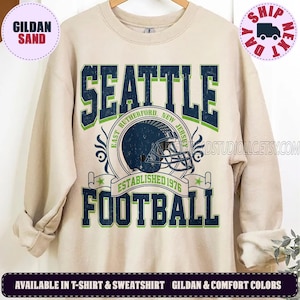 May include: A sand-colored crewneck sweatshirt featuring a graphic print with the text SEATTLE and FOOTBALL in dark blue with green outlines. The design includes a dark blue football helmet and the text EAST RUTHERFORD NEW JERSEY ESTABLISHED 1976.
