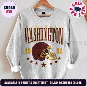 May include: A light gray crewneck sweatshirt featuring a distressed graphic of a maroon football helmet with a yellow facemask. The text reads WASHINGTON FOOTBALL 1932, accompanied by a row of maroon stars arranged in a semi-circle below the helmet.