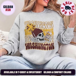 May include: A light gray crewneck sweatshirt featuring a graphic of a skull wearing a maroon beanie and holding a yellow pom-pom and a beverage can. The word WASHINGTON is printed in a wavy, retro-style font in shades of yellow and maroon.
