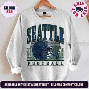 May include: A light gray crewneck sweatshirt featuring a distressed dark blue graphic of a football helmet centered between the numbers 19 and 76. The word SEATTLE is printed in large dark blue letters above the helmet, with FOOTBALL below.