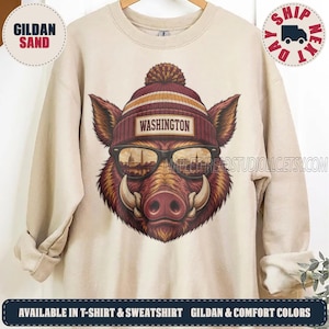 May include: A sand-colored crewneck sweatshirt featuring a graphic of a wild boar wearing a maroon and gold pom-pom beanie with the text WASHINGTON and sunglasses reflecting a city skyline. The boar has prominent tusks and brown fur.