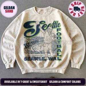 May include: A sand-colored crewneck sweatshirt featuring a vintage-style graphic of a football player in a jersey with the number 14, a stadium background, and the text Seattle Football Est. 1976 in dark blue and green ink on the front.