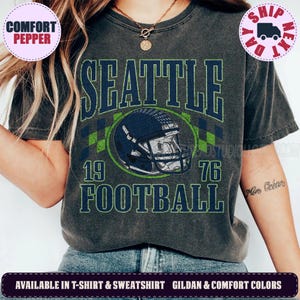 May include: A charcoal gray short-sleeve t-shirt featuring a graphic print with the text SEATTLE FOOTBALL 1976 and an illustration of a dark blue football helmet. The design includes a green and blue checkered pattern behind the helmet.
