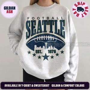 May include: A light gray crewneck sweatshirt featuring a dark blue graphic print. The design includes the text FOOTBALL SEATTLE, a football illustration containing a city skyline silhouette, the text EST. 1976, and a row of dark blue stars.