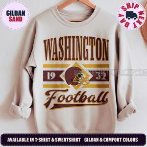 May include: A sand-colored crewneck sweatshirt featuring a graphic print with the text WASHINGTON, 1932, and Football. The design includes a maroon football helmet with a yellow facemask, set against horizontal yellow and maroon stripes.