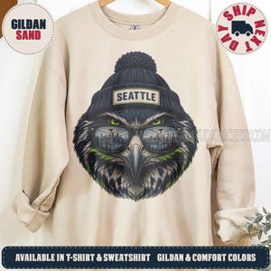 May include: A sand-colored crewneck sweatshirt featuring a graphic of an eagle wearing a dark blue knit beanie with a pom-pom and a patch that reads SEATTLE. The eagle wears sunglasses reflecting a city skyline with a tall tower.