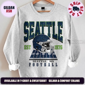 May include: A light grey crewneck sweatshirt featuring a dark blue football helmet graphic centred above a football field design. The text reads SEATTLE EST 1976 SEATTLE, WA FOOTBALL in dark blue and lime green lettering with a distressed texture.