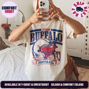 May include: An ivory short-sleeve t-shirt featuring a graphic print of a football helmet in red and blue. The text reads 1960 BUFFALO FOOTBALL in a distressed blue font. The design is centered on the chest of the shirt, which has a crew neckline.
