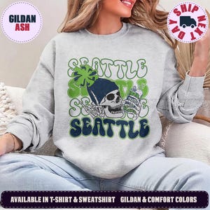 May include: A light gray crewneck sweatshirt featuring a graphic of a skeleton wearing a dark blue beanie and holding a green can. The text SEATTLE is repeated in a wavy font in green and dark blue. A green pom-pom sits atop the beanie.