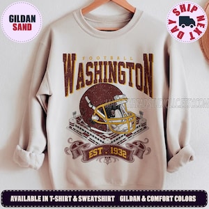 May include: A sand-colored crewneck sweatshirt featuring a graphic print of a maroon football helmet with a yellow facemask, a stadium illustration, and the text WASHINGTON FOOTBALL EST. 1932 in maroon and yellow lettering on the front.